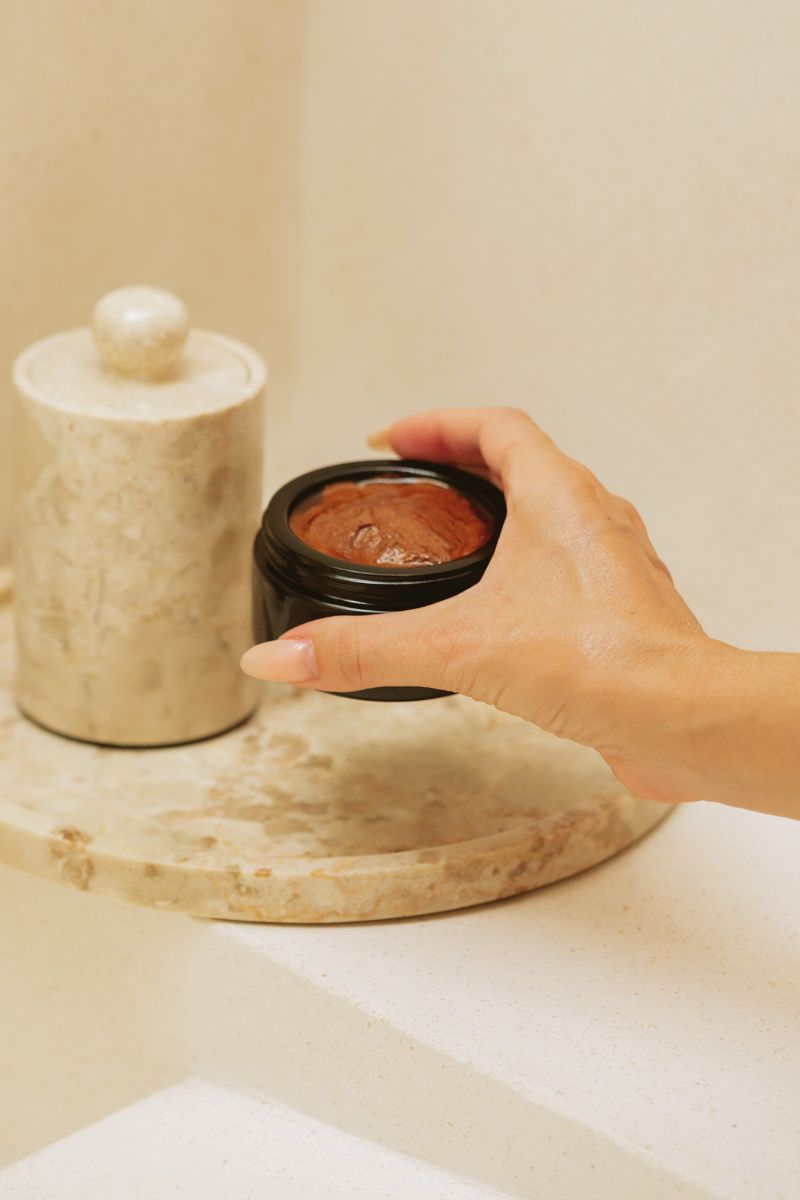 Hand picking up black jar of earthy colored face mask in stone bathroom.