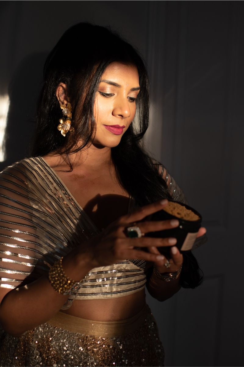 Woman dressed in bright gold and silver clothing with light illuminating her face and glowing skin looking at the instructions for UMM Skincare's body scrub.