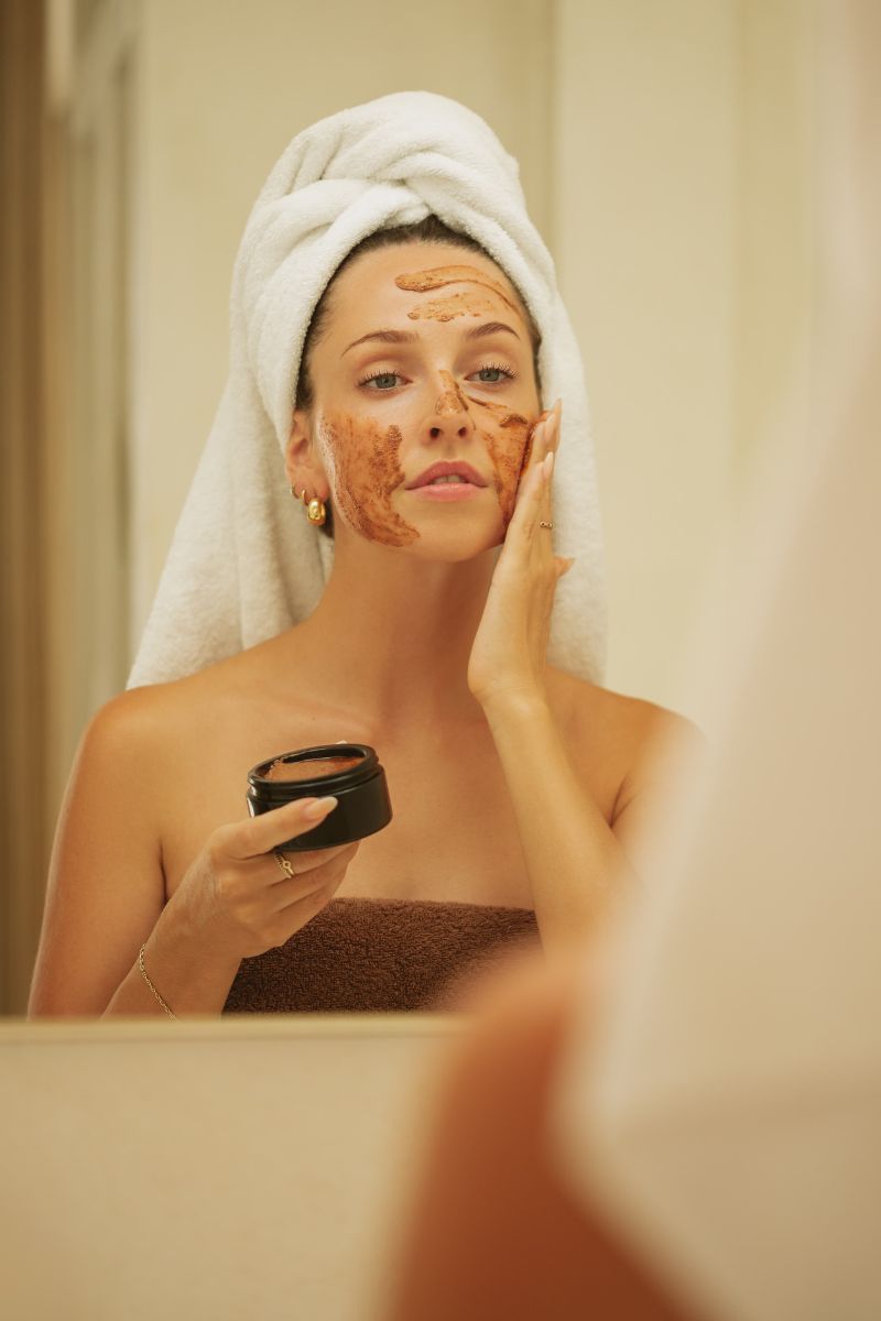 Woman with a towel wrapped around her head applying a clay face mask in front of a mirror, holding a jar of the skincare product, emphasizing a relaxing self-care ritual.